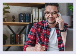A man with grey hair and a plaid shirt sitting before a laptop, on his cell phone. He is taking notes.